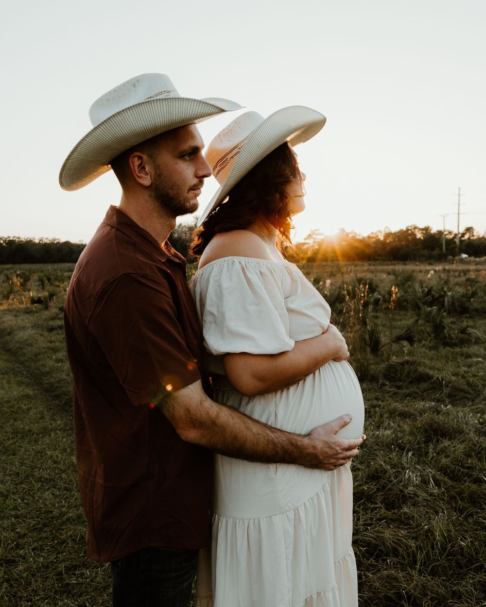 Couple in cowboy hats embracing at sunset in a field, pregnant woman in white dress holding her baby bump.