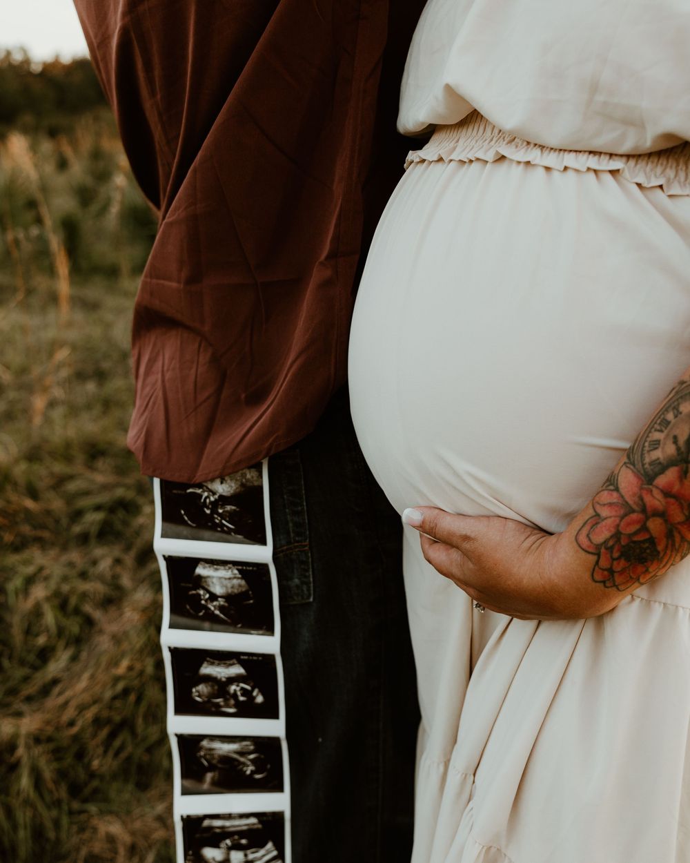 Pregnant woman in white dress with tattoos and partner holding ultrasound strip photos outdoors.