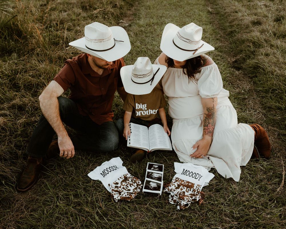Family pregnancy announcement with cowboy hats, ultrasound photos, and big brother shirt on grass.
