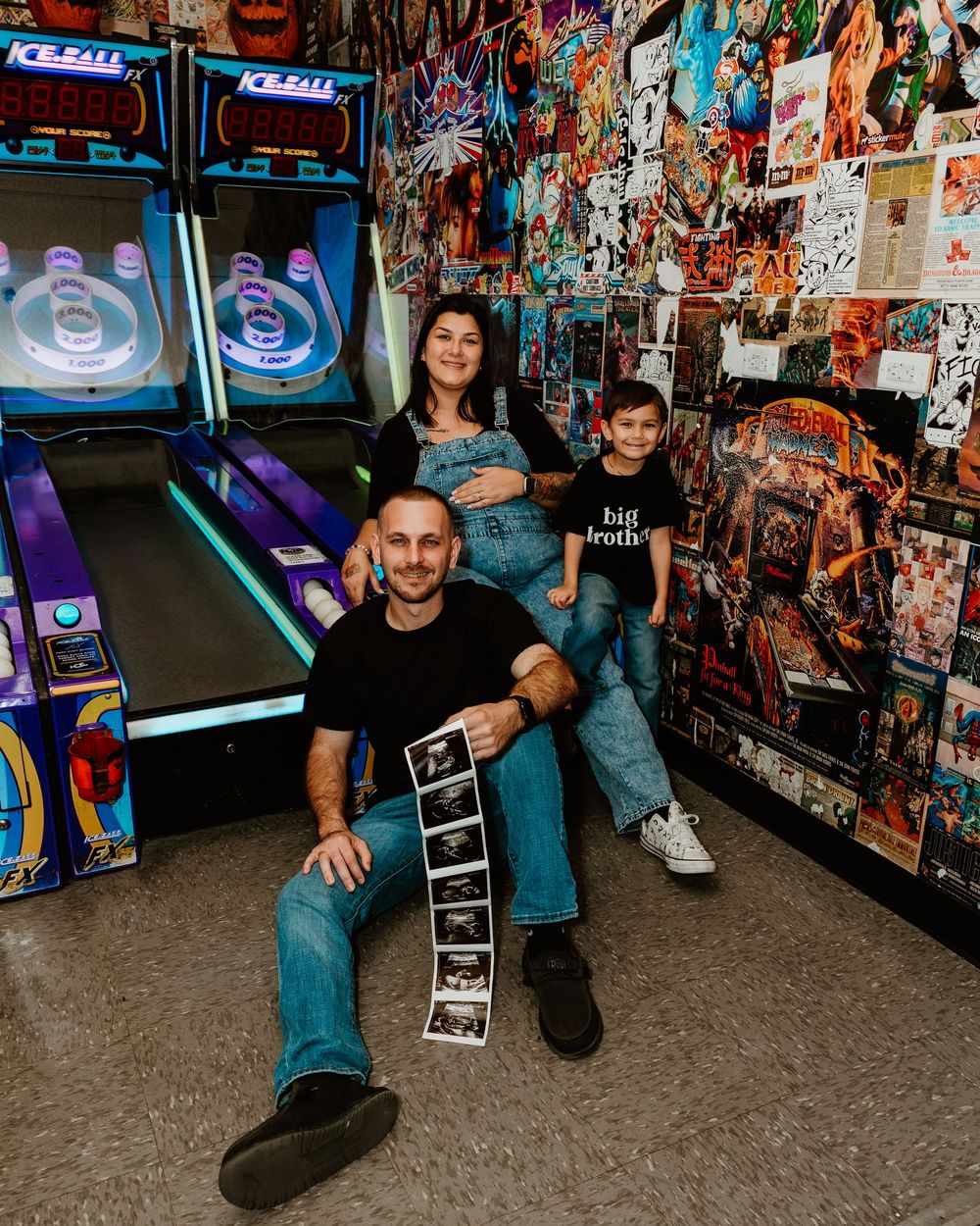 Family of three poses in arcade with skee-ball machines, holding ultrasound photos, child wearing big brother shirt.