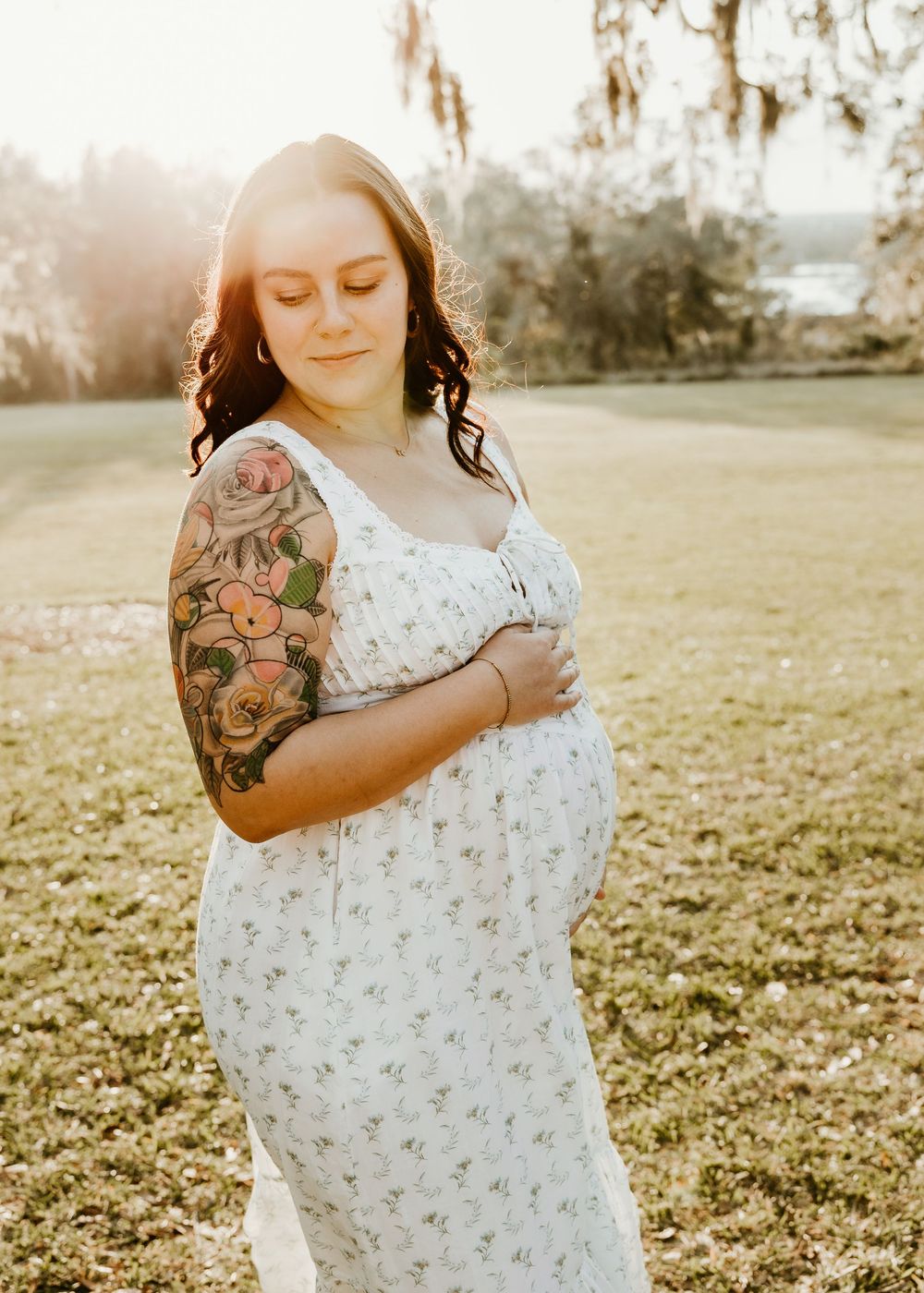Woman in a white floral dress shows off her baby bump and colorful arm tattoo in golden sunlight.