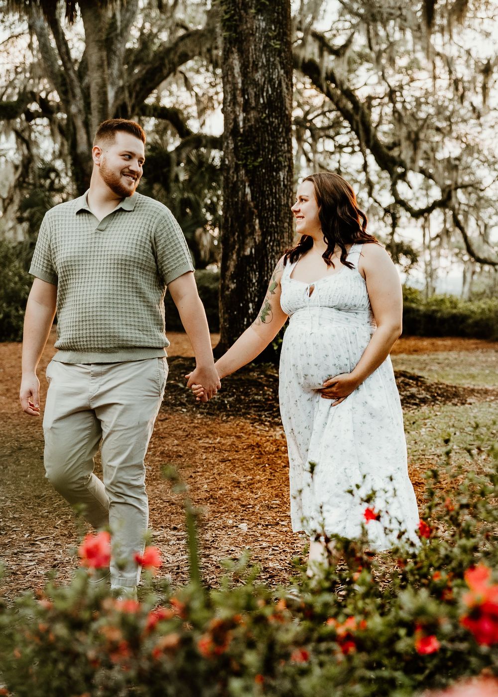 Couple walking hand in hand through a garden with red flowers and Spanish moss trees.