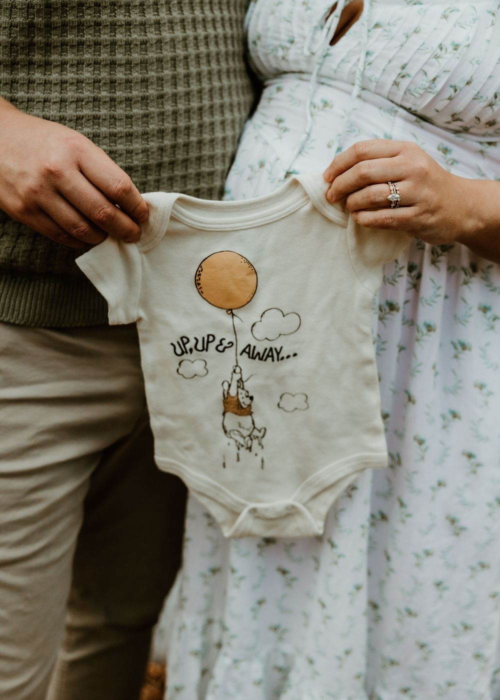 Couple holding up a white baby onesie with a cute balloon and cloud design.