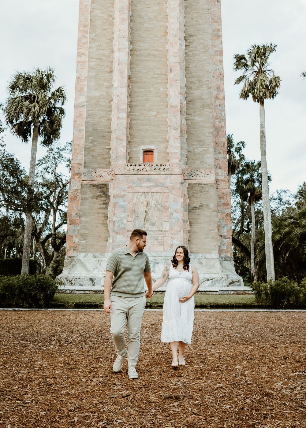 A couple stands in front of a tall stone tower with palm trees in the background.