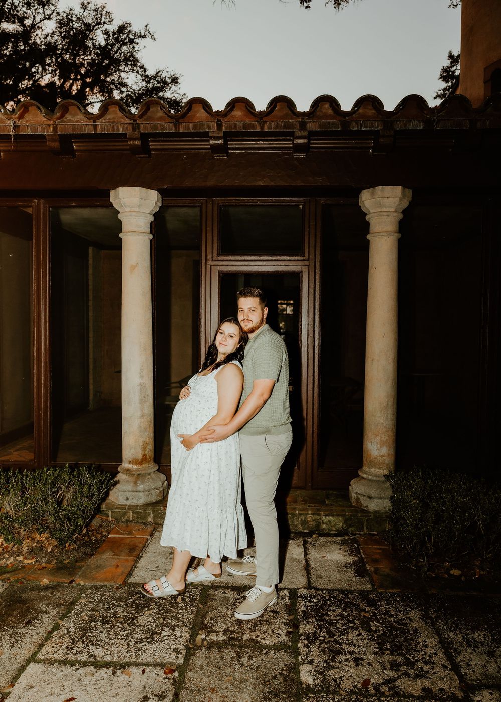 Couple posing between classical columns at sunset during maternity photo session.