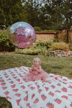 Baby wearing pink tutu dress sitting on a polka dot blanket in a sunny backyard garden setting.