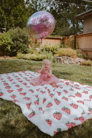 A pink and white strawberry-themed first birthday party setup in a backyard with a balloon and blanket.