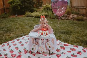 Baby in white dress sits in high chair with pink balloon and watermelon patterned blanket for first birthday celebration.