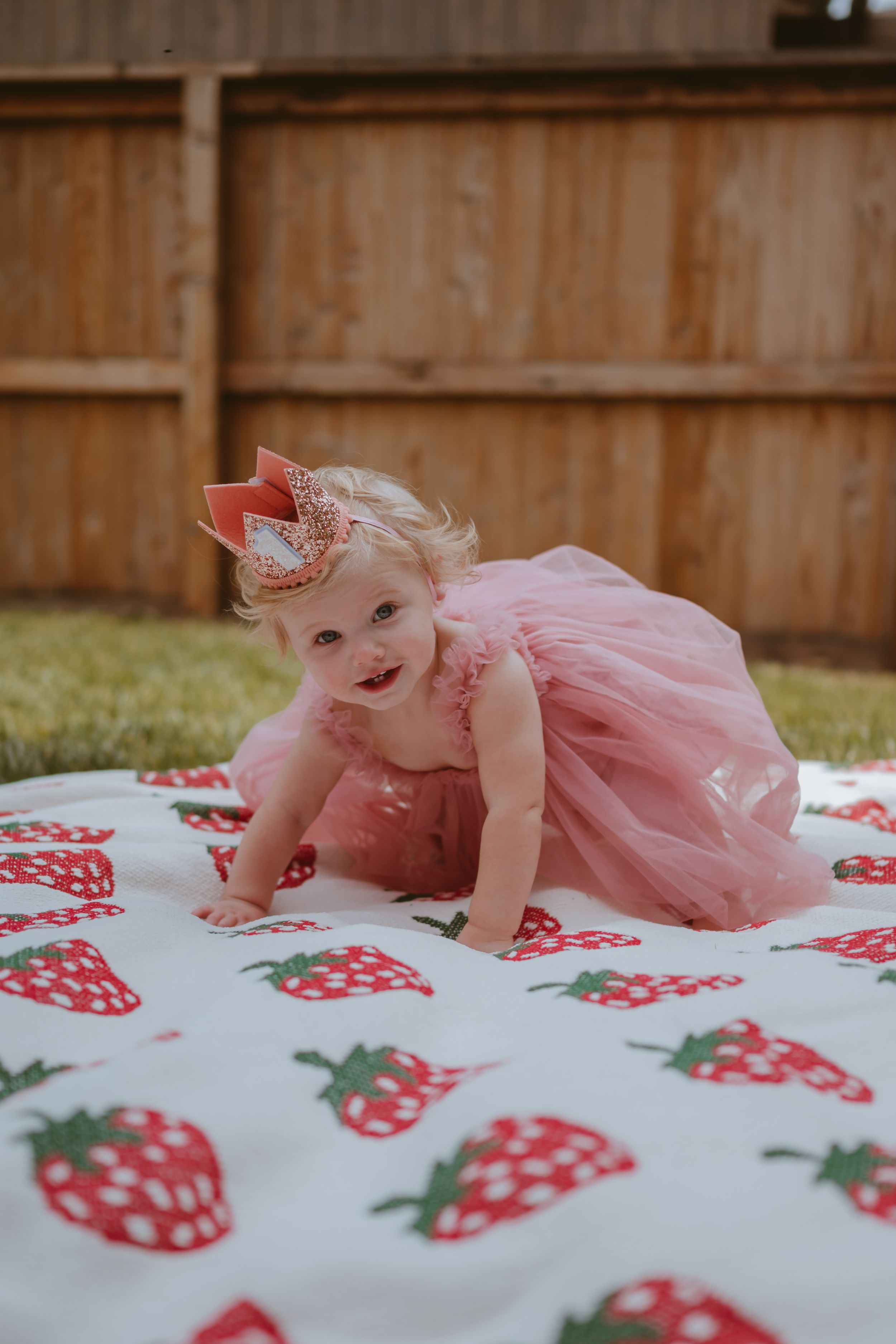 A small child in a pink tulle dress crawls on a white blanket with red strawberry pattern.