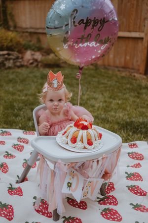 Baby in pink dress sits next to strawberry-themed birthday cake with balloons at outdoor first birthday celebration.