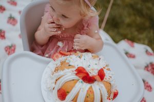 A toddler in pink sits before a festive bundt cake decorated with strawberries and orange slices on a white plate.