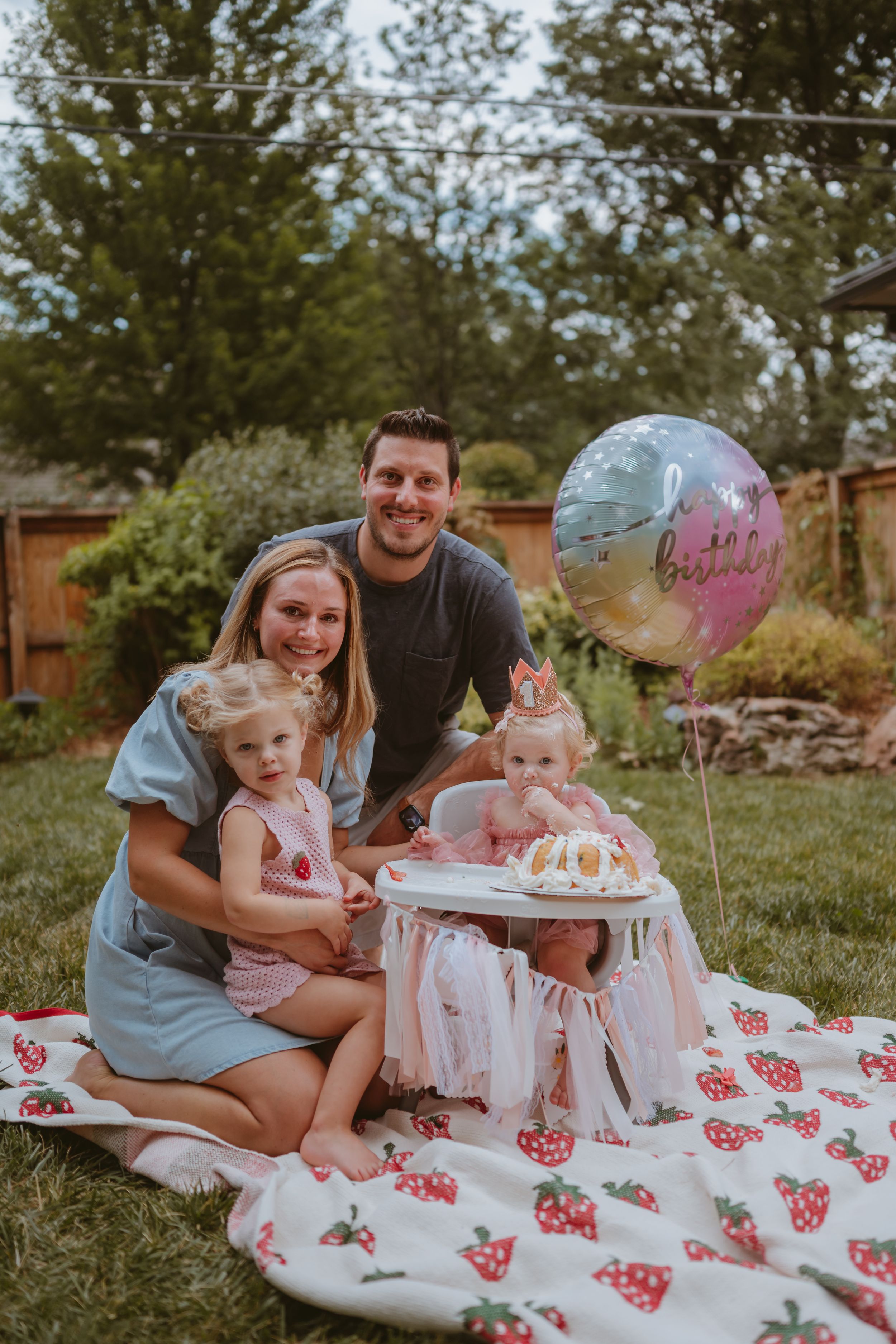 Family enjoying outdoor birthday celebration with cake on strawberry print blanket in backyard garden setting.
