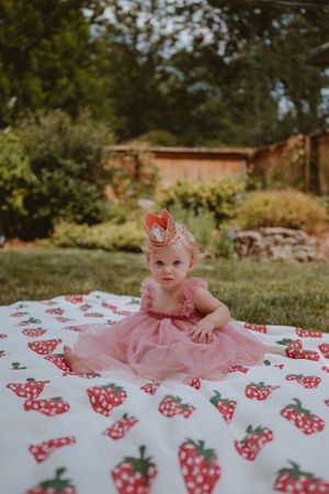 Baby in pink tulle dress lying on white blanket with red strawberry pattern in outdoor garden setting.