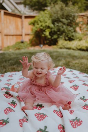 Happy baby wearing pink tulle dress sits on white blanket with strawberry pattern in sunny backyard garden.