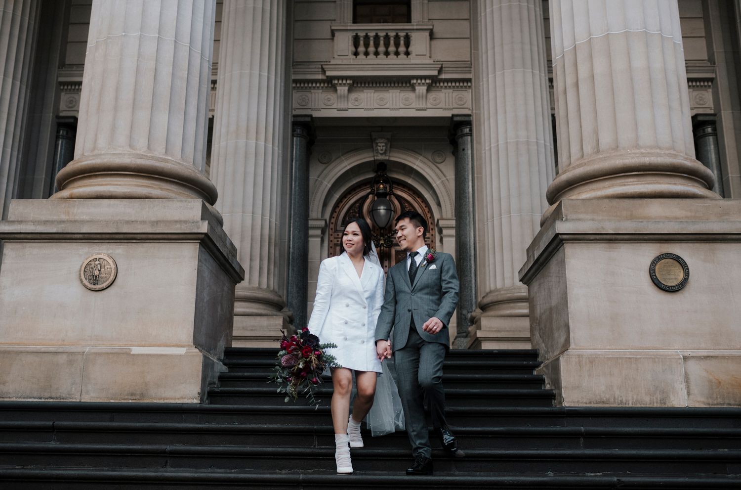 A couple descends stone steps at an elegant building entrance with classical columns and architectural details.