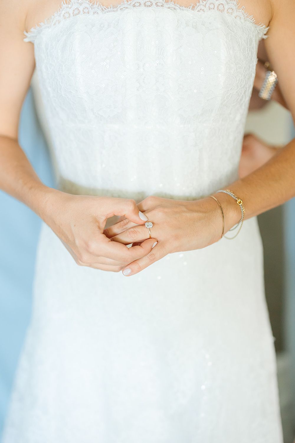 Close-up of hands with delicate jewelry against a white lace wedding dress, showing elegant bridal details.