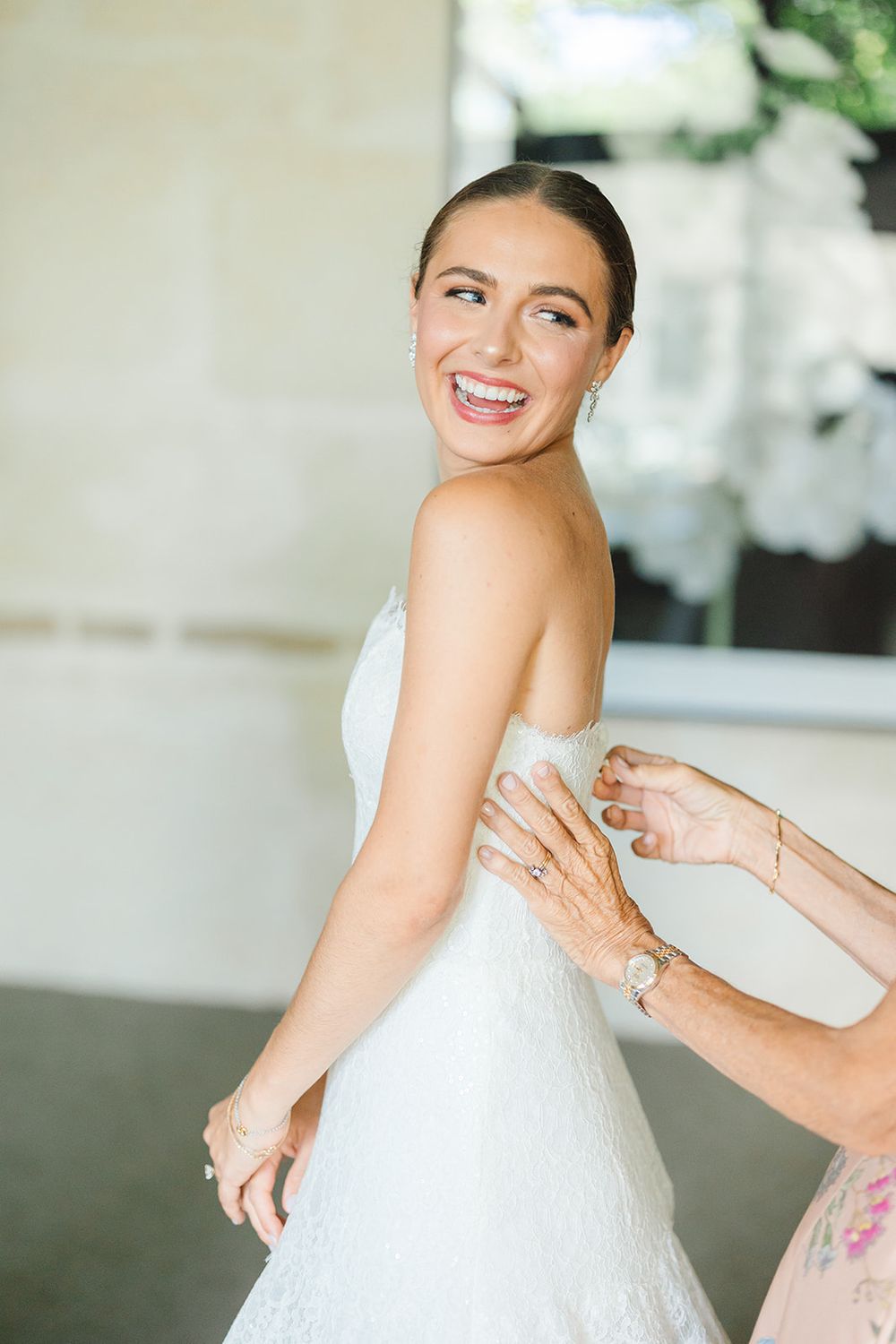 A bride wearing a white wedding dress smiles joyfully while getting help with final dress adjustments.