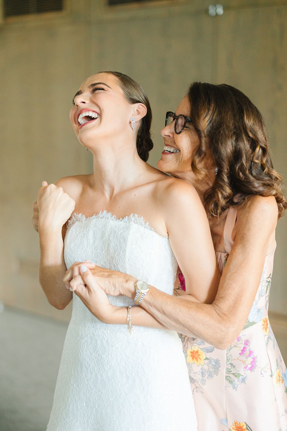 Two people in white dresses share a joyful, candid moment while laughing together.