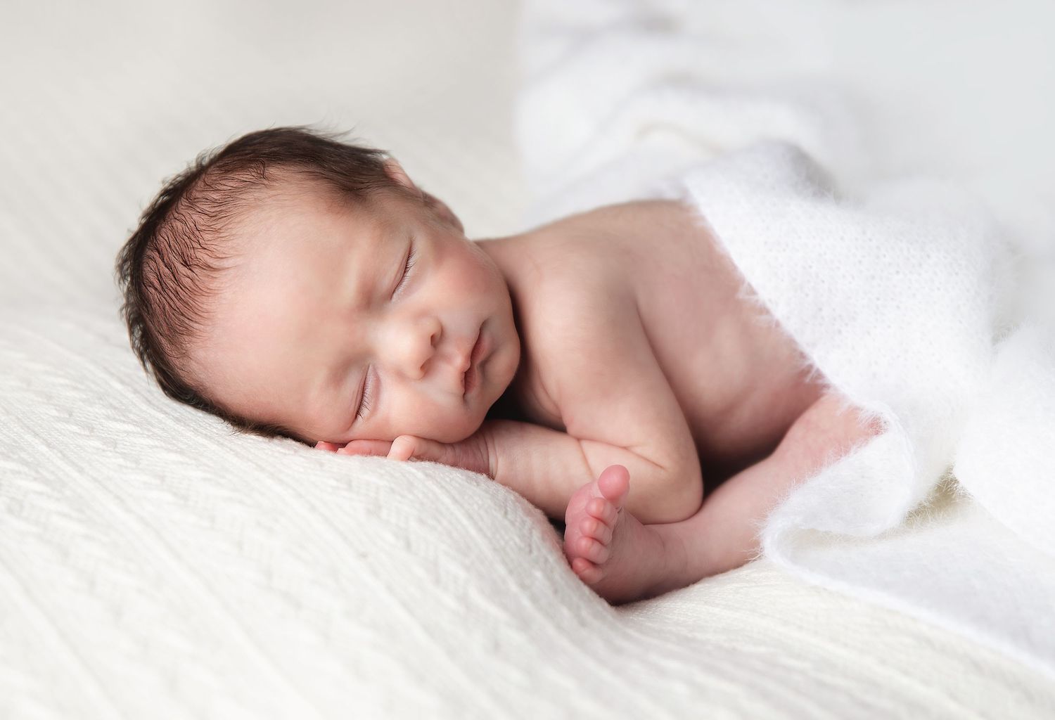 Peaceful sleeping newborn baby wrapped in soft white blanket on light background. During a newborn session in Bexley