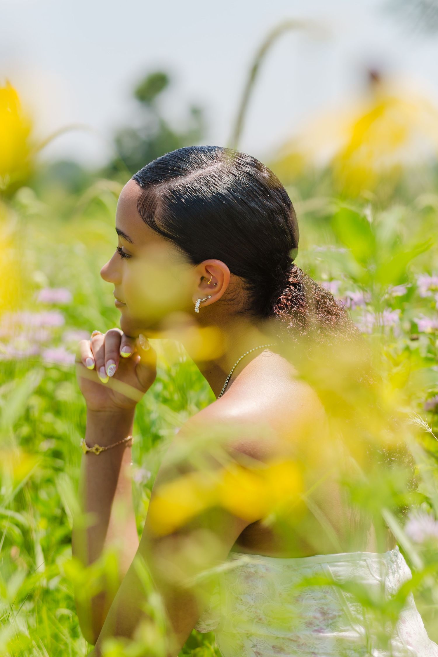 A young woman sits thoughtfully in a garden surrounded by yellow flowers and greenery, photographed in a dreamy side profile.