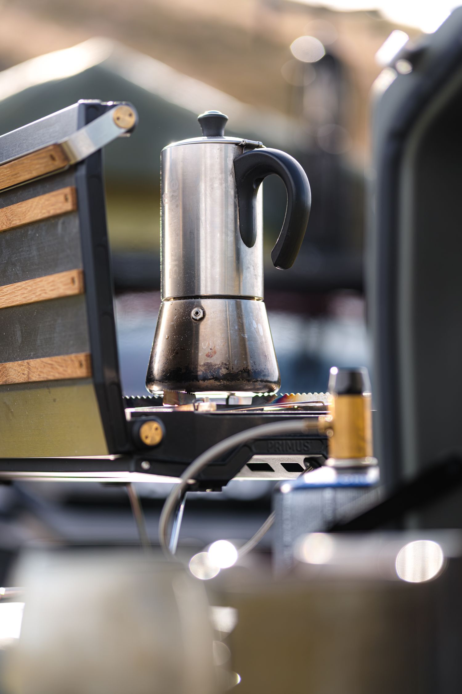 A stainless steel stovetop espresso maker sits on a portable camping stove in an outdoor setting.