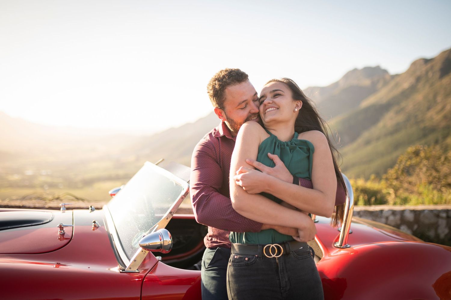 A couple shares tender moments in a red vintage car against a mountainous backdrop at sunset.