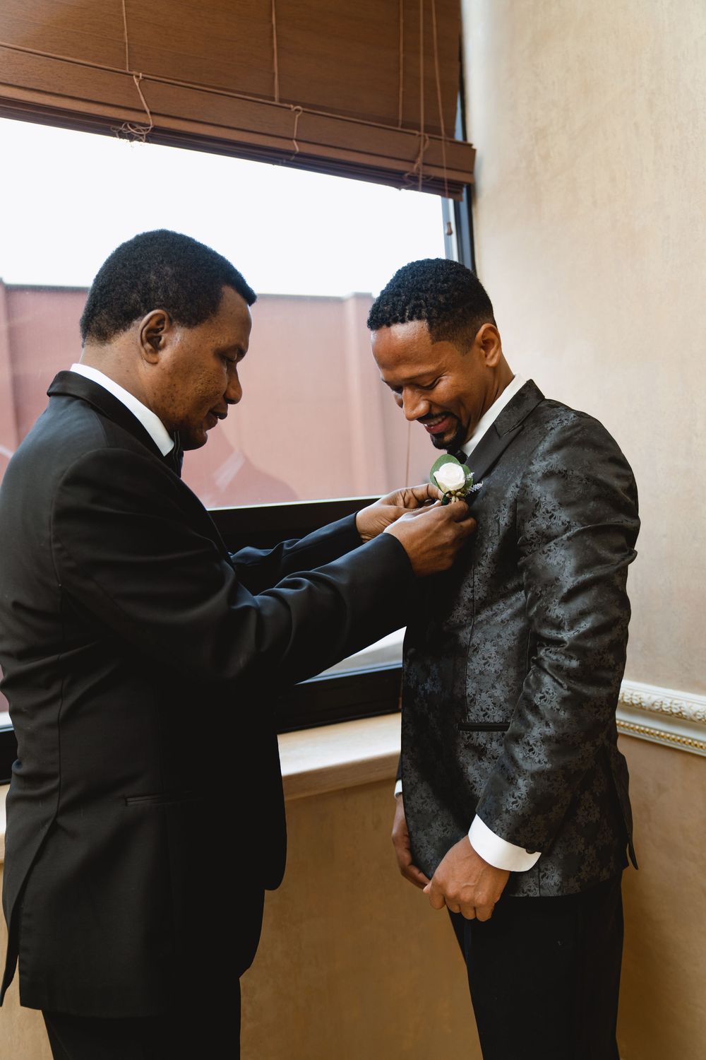 A touching moment as a man helps pin a white boutonniere to another's dark textured jacket.