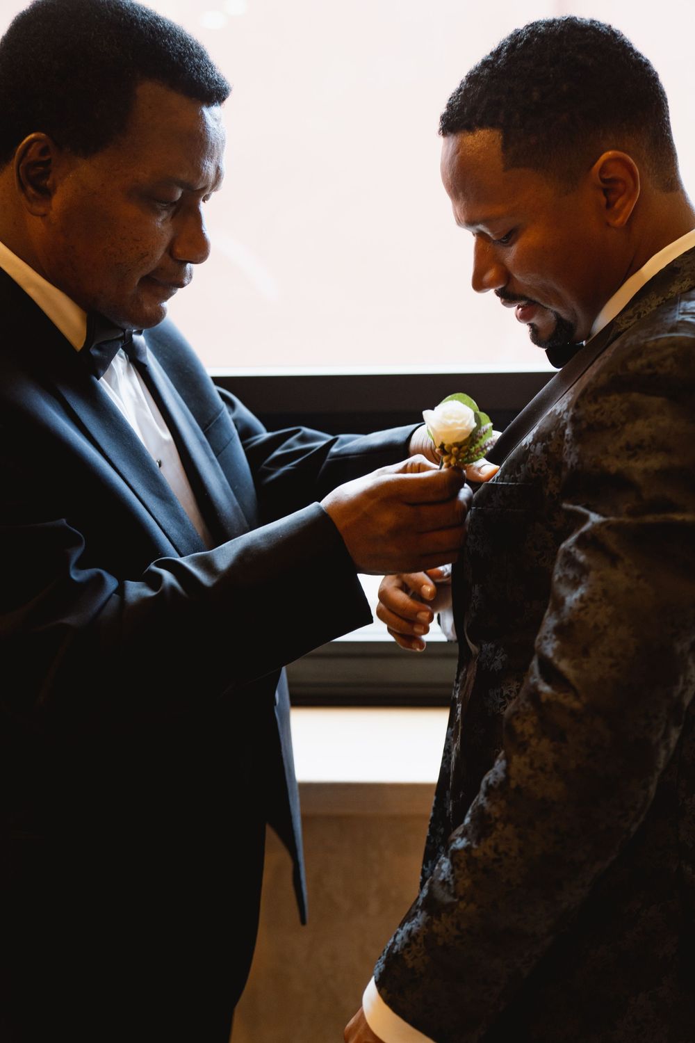 Two men in formal attire share an intimate moment as one helps adjust the other's boutonniere with care.