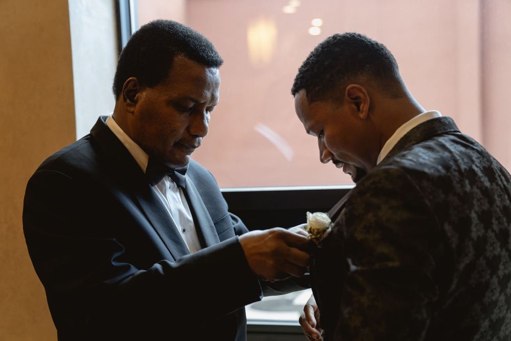 A candid shot captures two men during a formal dressing ritual with one adjusting the other's boutonniere.
