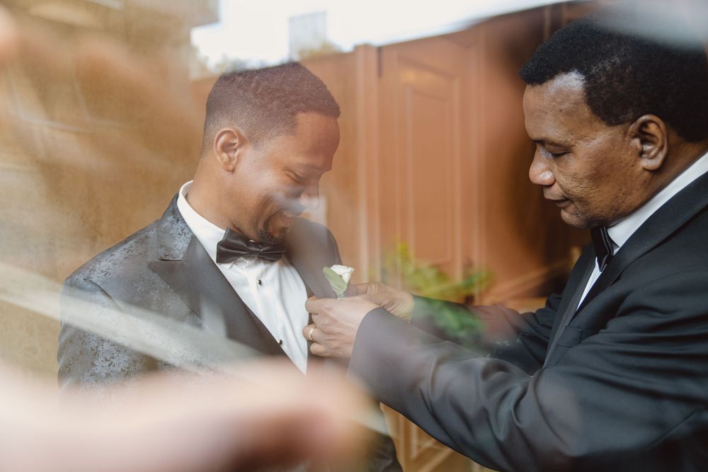 Through glass, two men in bow ties share a moment as one assists with formal attire preparations.