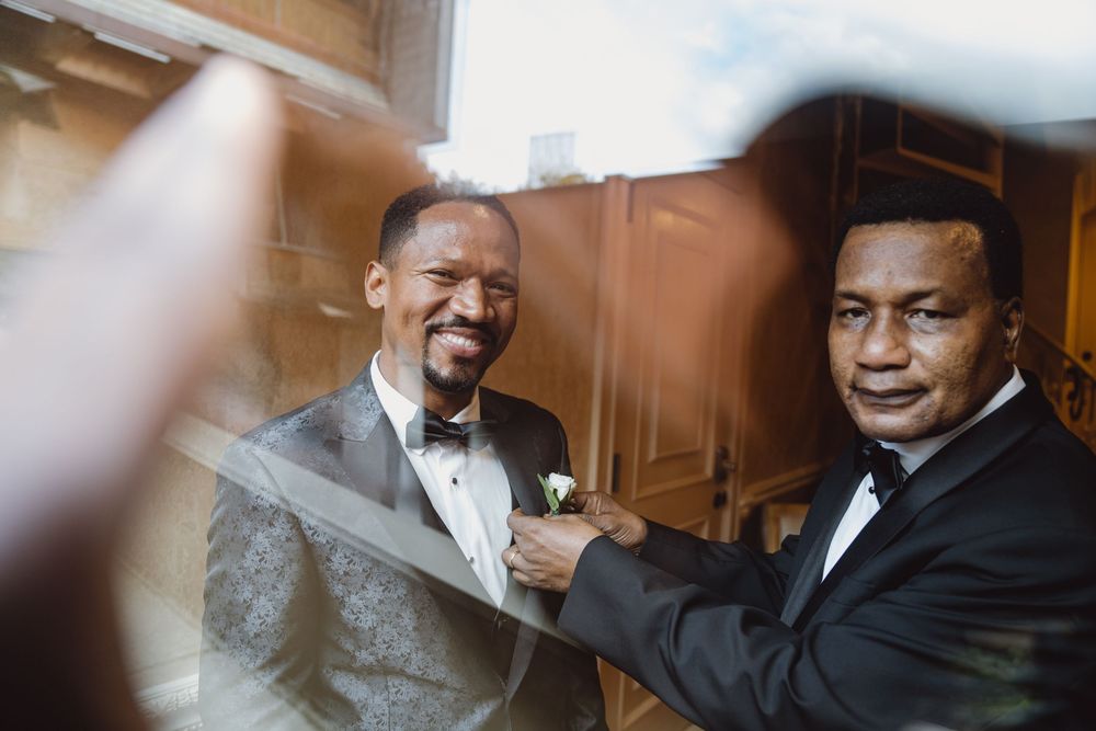 Two men in formal wear share a joyful moment while adjusting each other's attire.