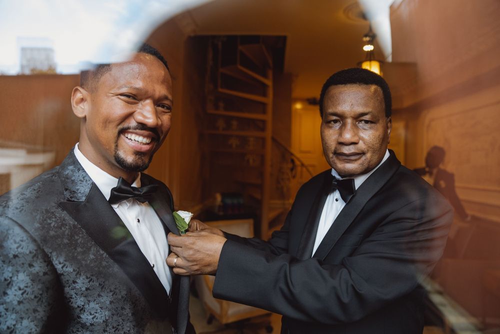 Two men in tuxedos laugh while helping each other prepare for a formal event.