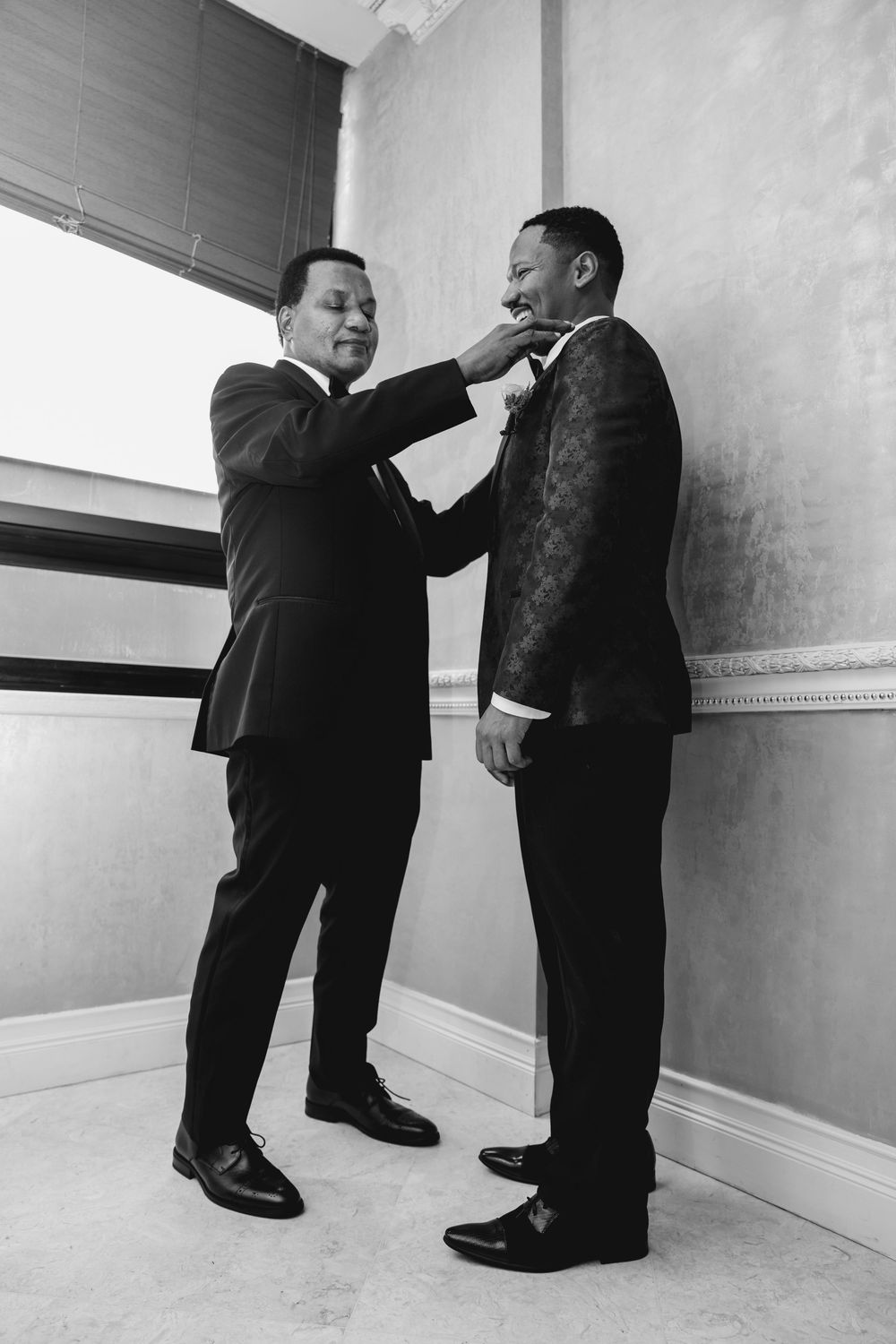 A black and white photo shows two men in suits during a pre-event grooming moment.
