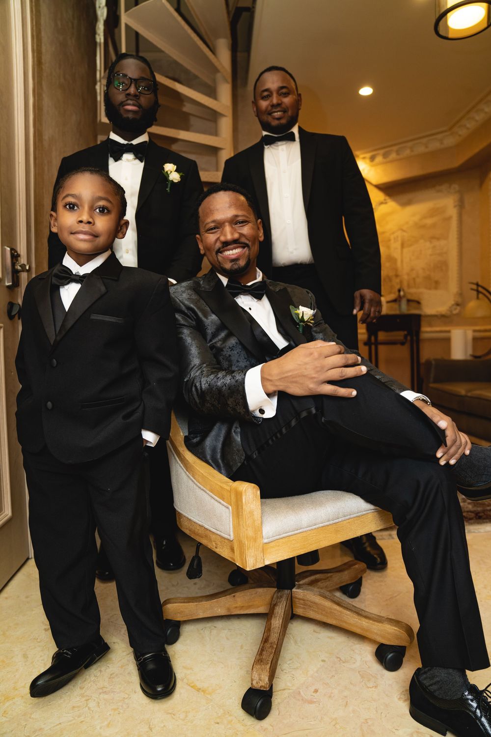 A formal group portrait shows four people in tuxedos gathered around a chair in an elegant setting.