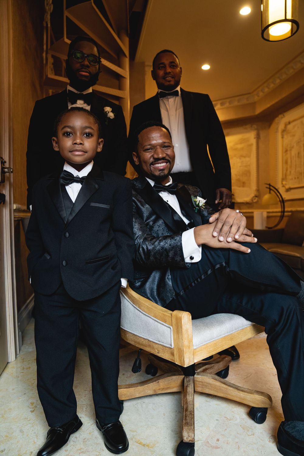 A group of men in black tuxedos sitting casually on a cream-colored chair during wedding preparations.