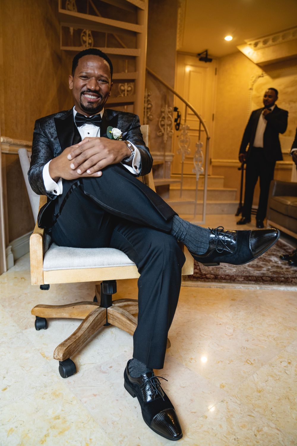 Wedding party member lounging in office chair wearing black tuxedo with white boutonniere.