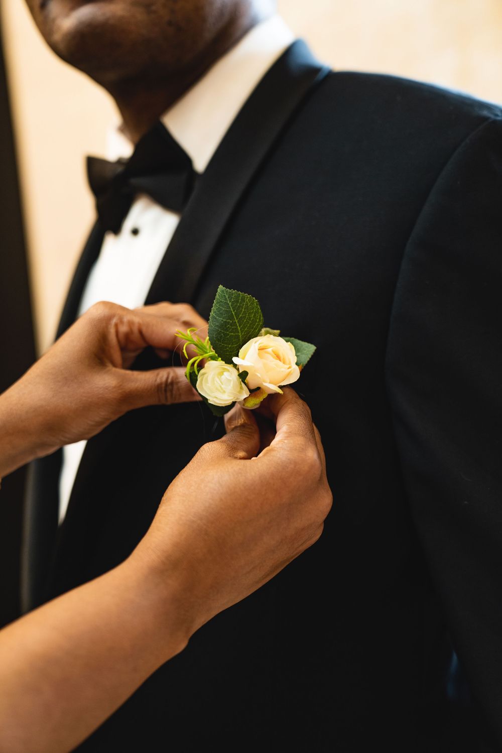 Close-up of hands pinning white rose boutonniere on black tuxedo lapel.