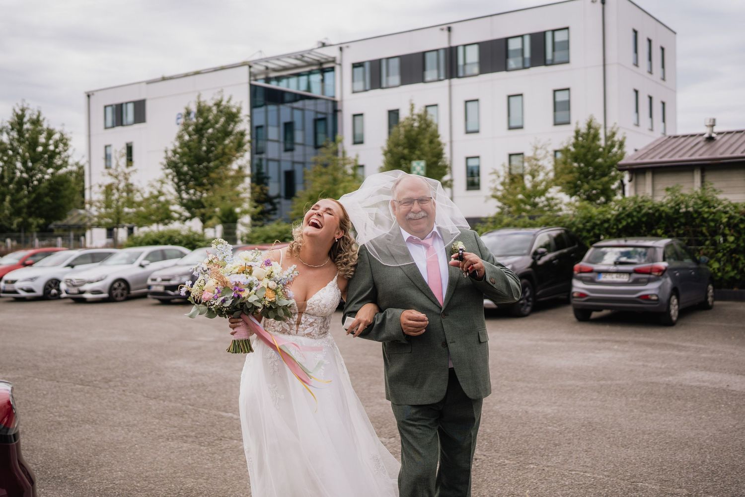 Wedding couple shares a candid moment in a parking lot with modern buildings in the background.