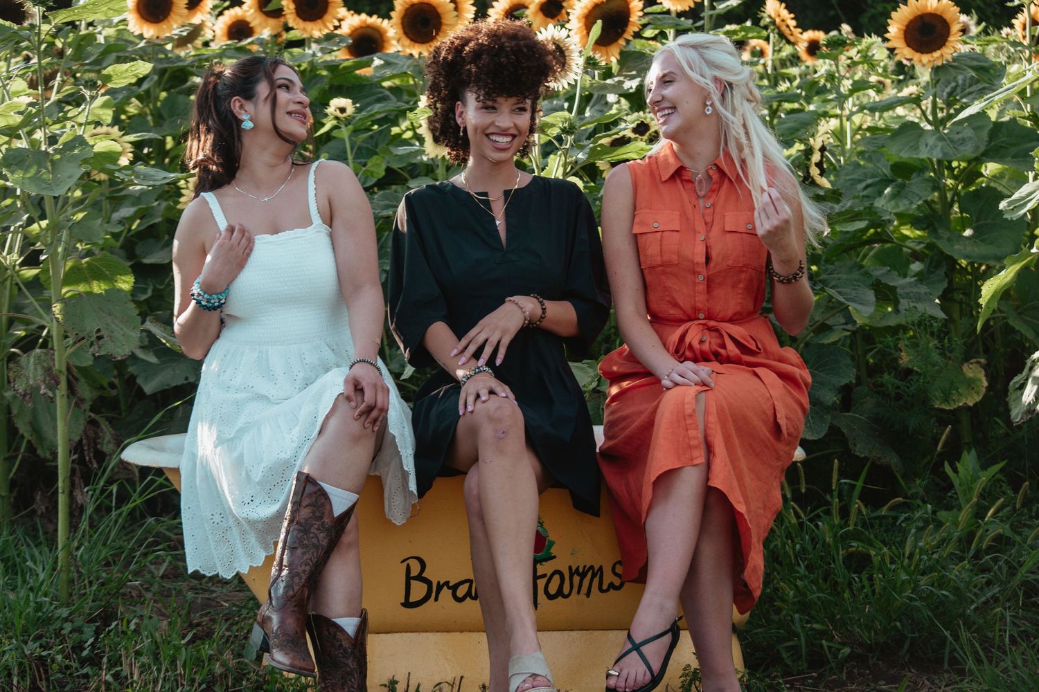 Three friends in summer dresses share a laugh while sitting on a wooden bench in front of a blooming sunflower field.