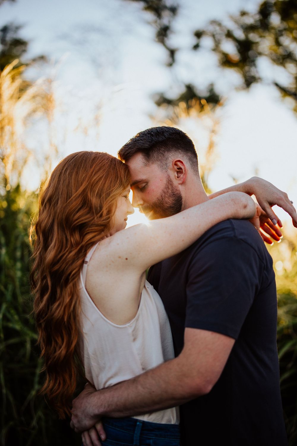 A romantic couple embraces in front of golden sunlit grass during a warm sunset photoshoot.