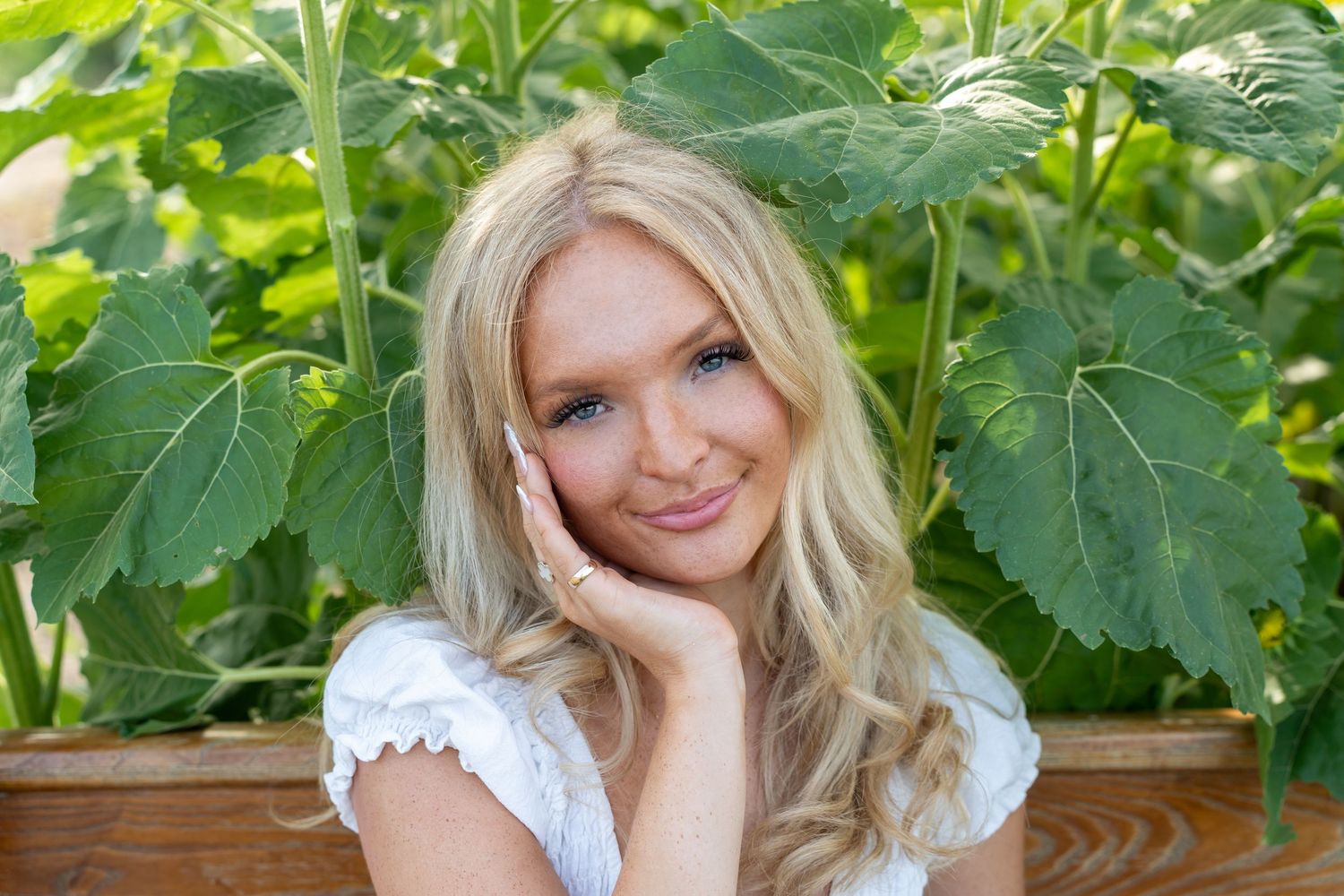 A woman stands smiling in a garden wearing a flowing white dress surrounded by lush green foliage.
