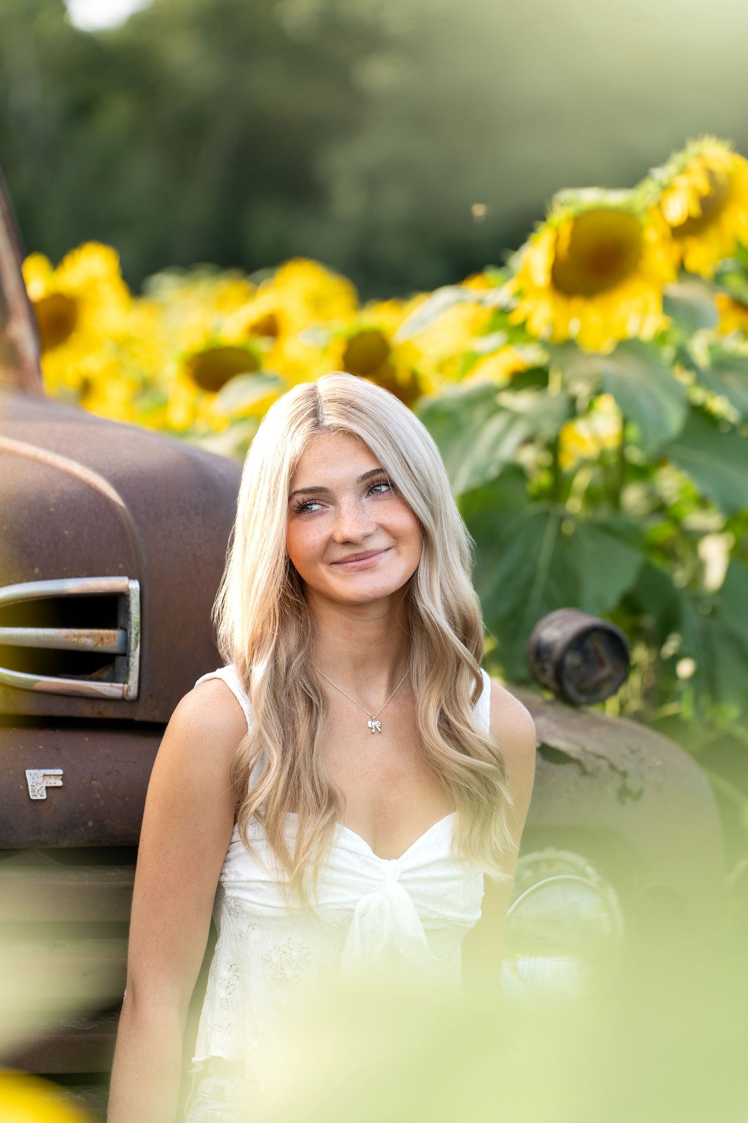 A white dress stands out against vibrant yellow sunflowers and a rustic truck in a summer portrait setting.