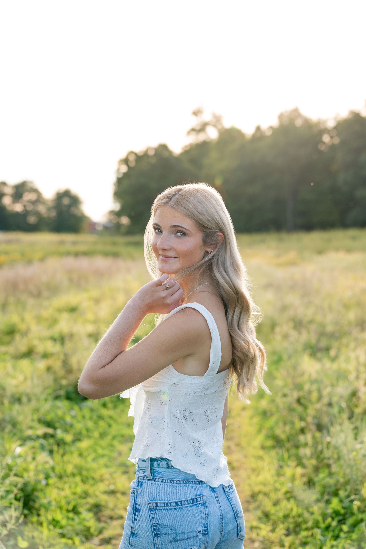 Young woman in denim overalls and white tank top poses playfully in a sunlit meadow during golden hour.