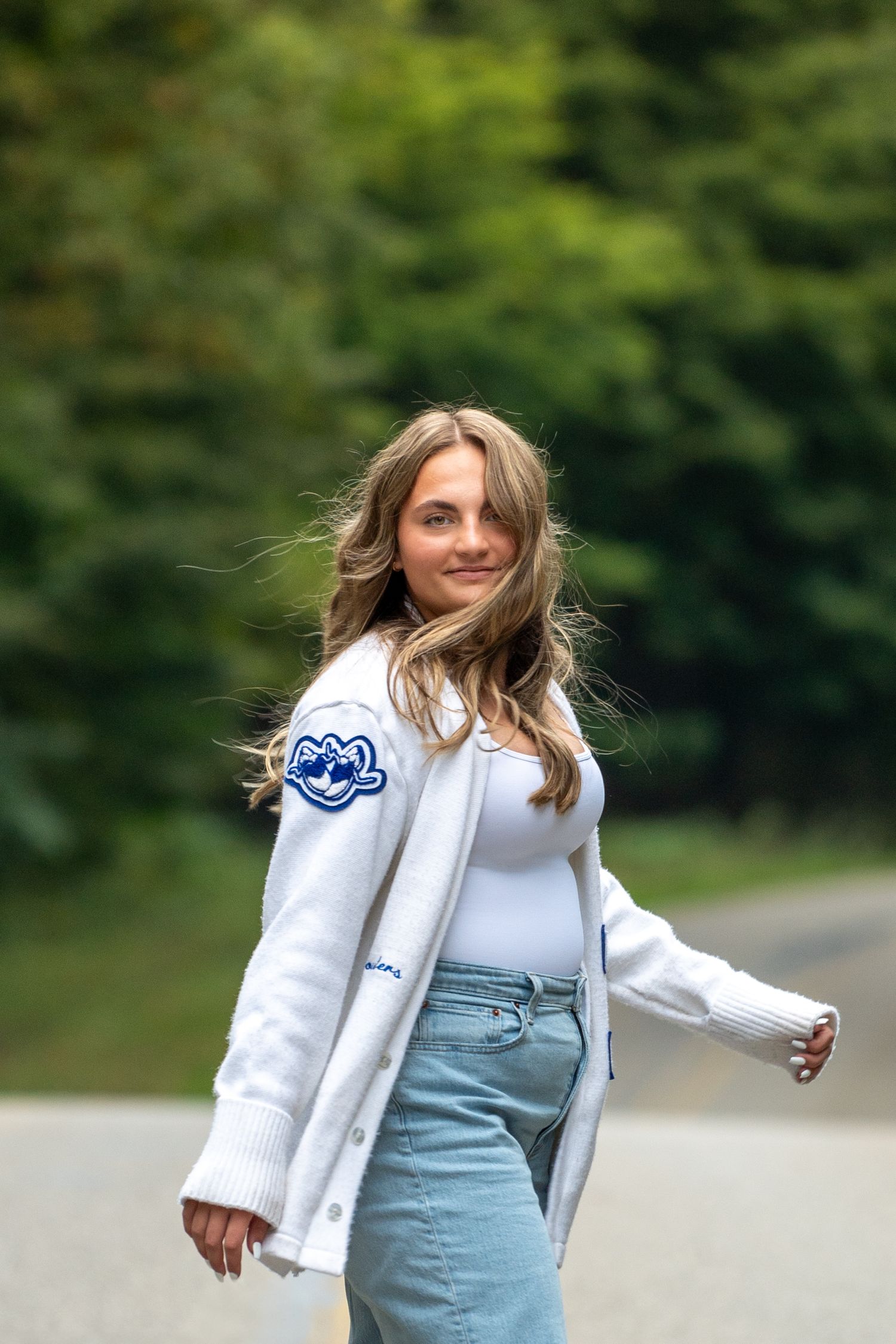 Fashion model in white blazer and light blue dress poses outdoors against blurred green foliage background.
