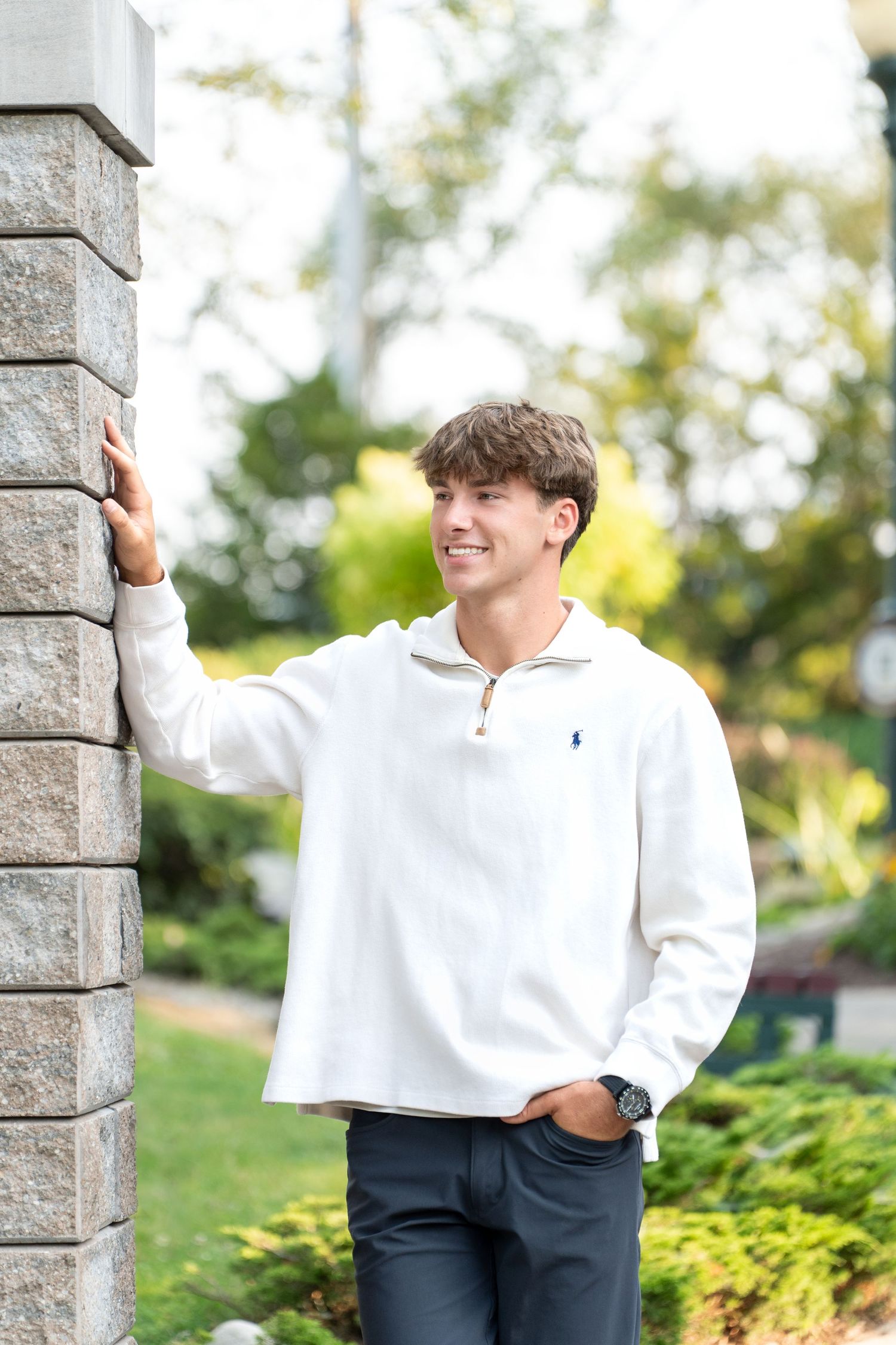 Smiling young man in white Polo Ralph Lauren quarter-zip leaning against a stone pillar outdoors.
