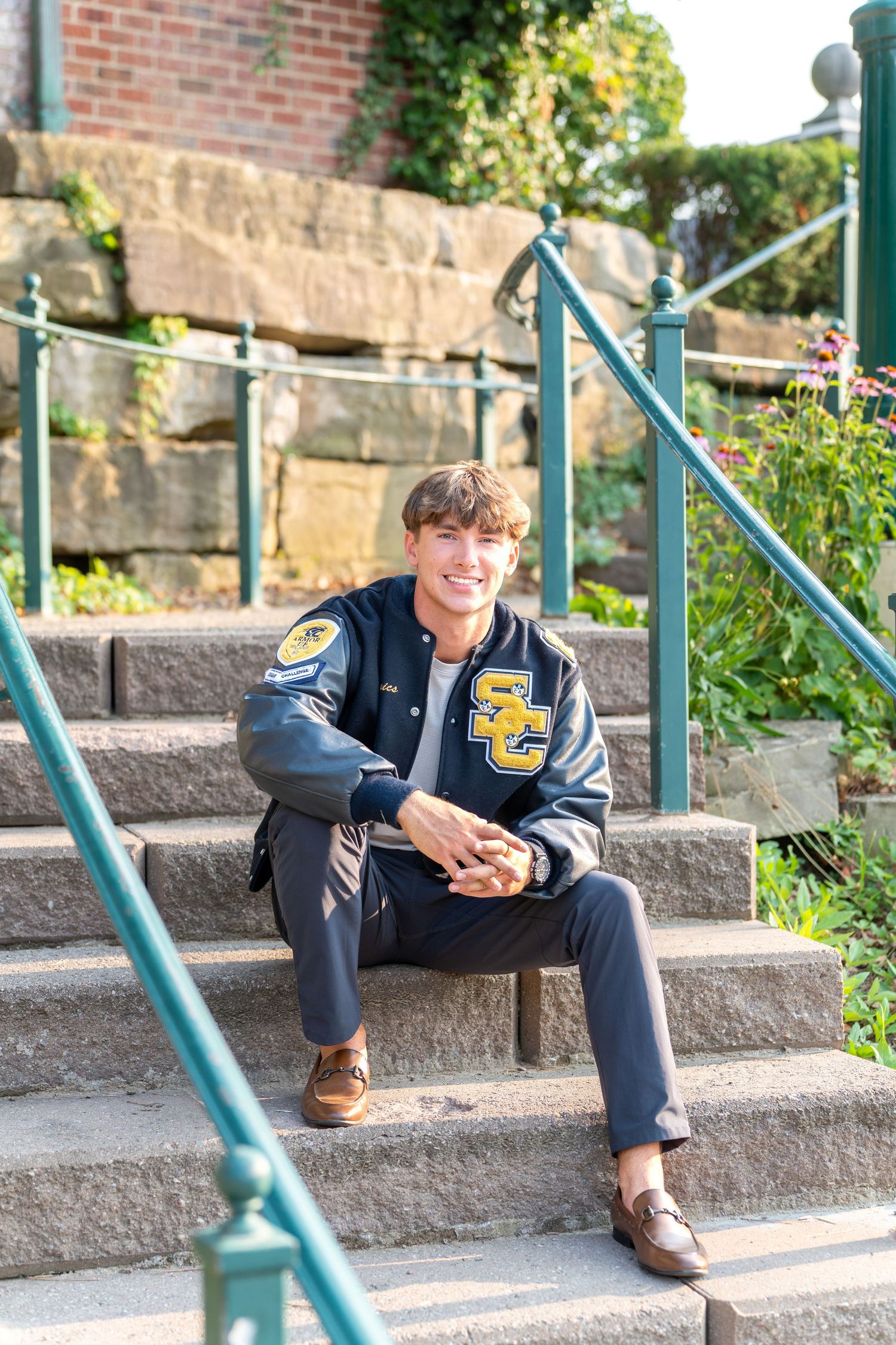 Smiling young man in SC letterman jacket sitting on stone steps outdoors in a sunny garden setting.
