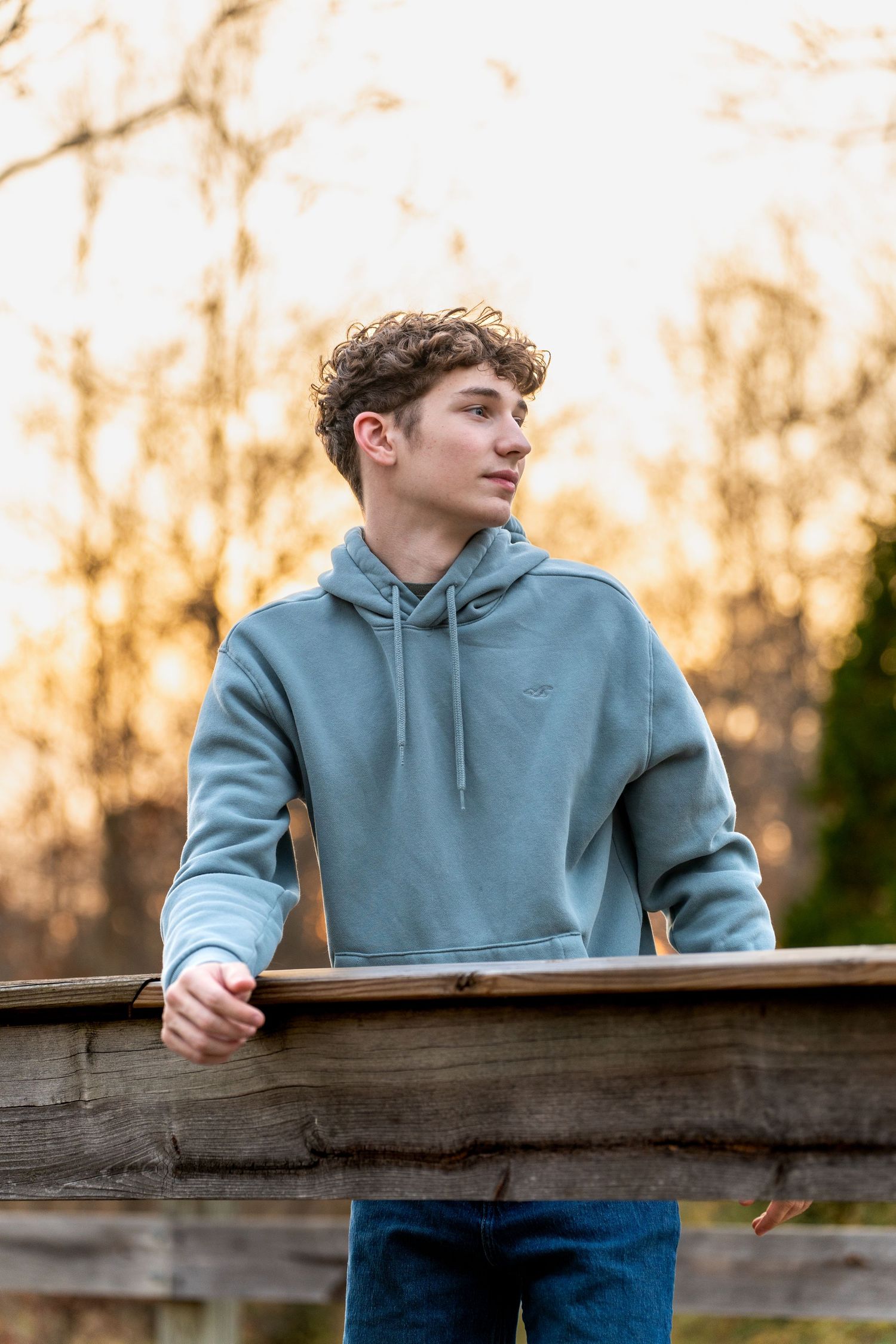 Person in a blue hoodie sweatshirt leans against a wooden fence during golden sunset light.