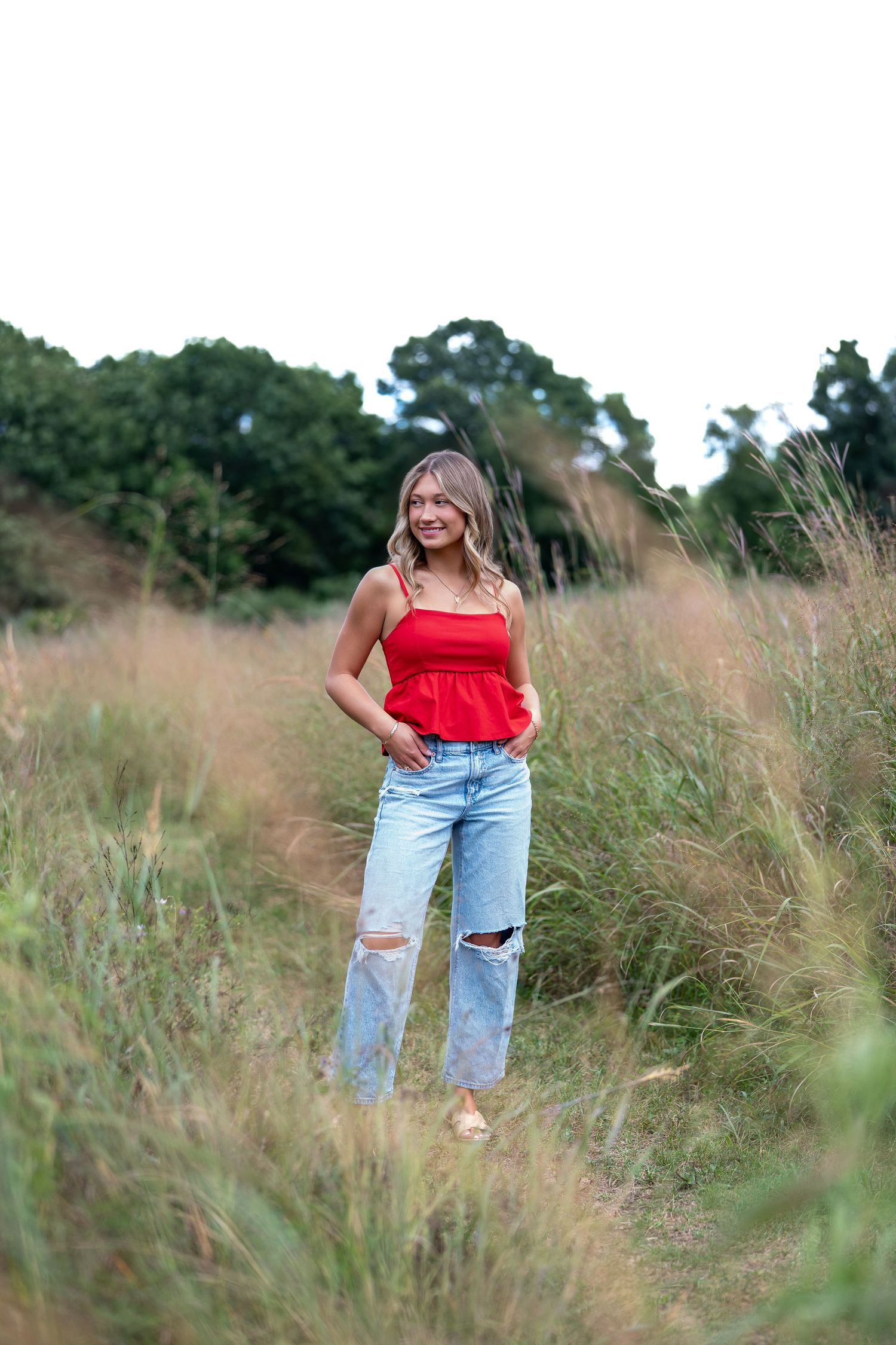 A person in a red off-shoulder top and distressed jeans poses in a field of tall grass during golden hour.