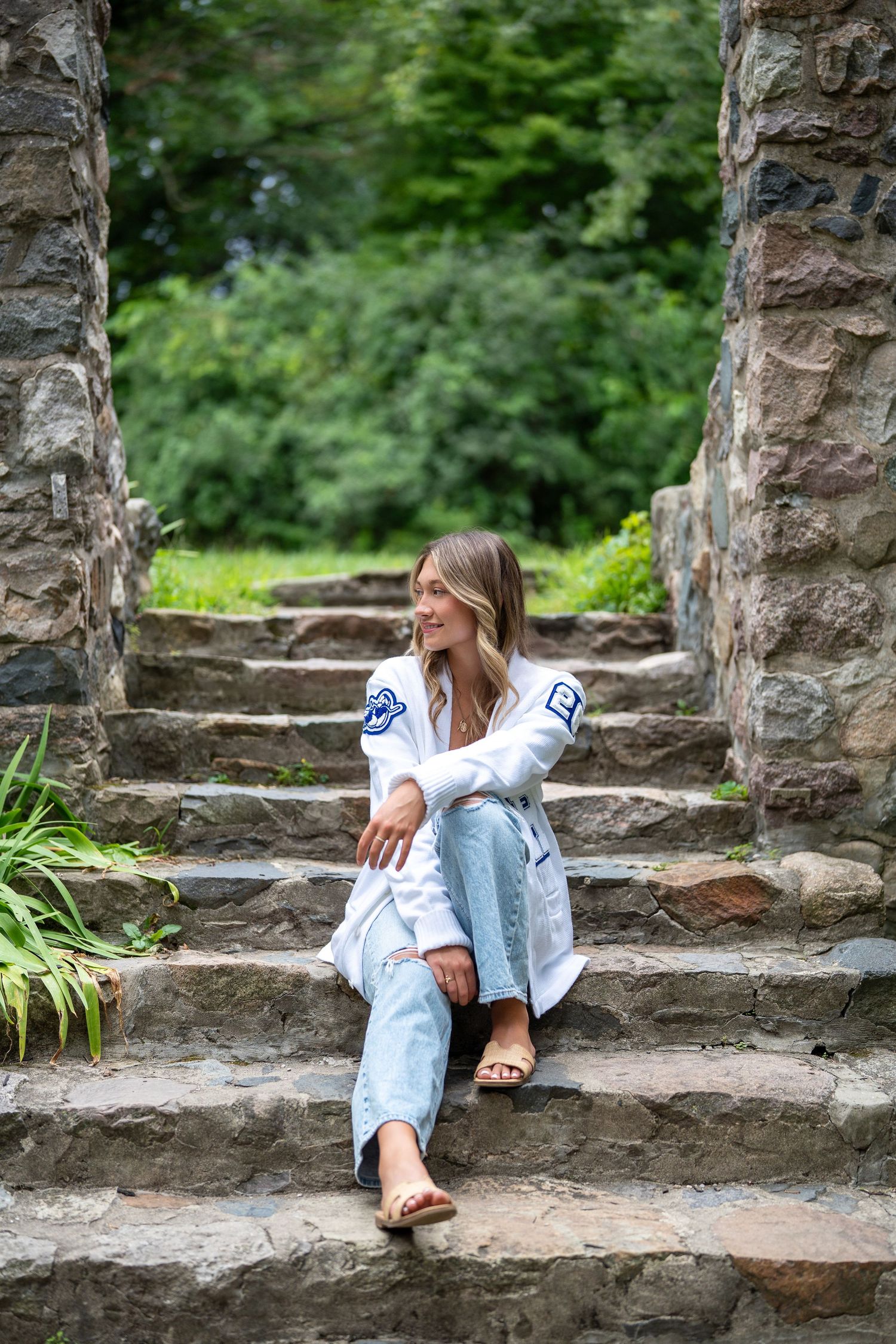 A person in a white embroidered top and light jeans sits casually on rustic stone steps surrounded by greenery.