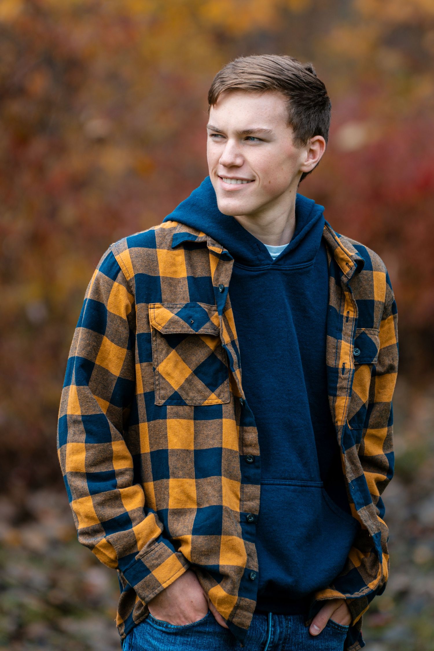 A person wearing a yellow and black plaid flannel shirt over a navy hoodie poses outdoors against autumn foliage.